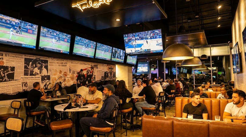 A lively sports bar with patrons watching multiple games on large screens, featuring a "Tailgate" neon sign.