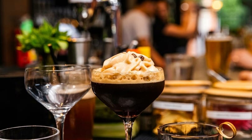 Dark cocktail with frothy cream and coffee beans on top, with blurred restaurant background.